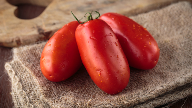 A trio of fresh, red San Marzano tomatoes sitting on some burlap with a cutting board in the background