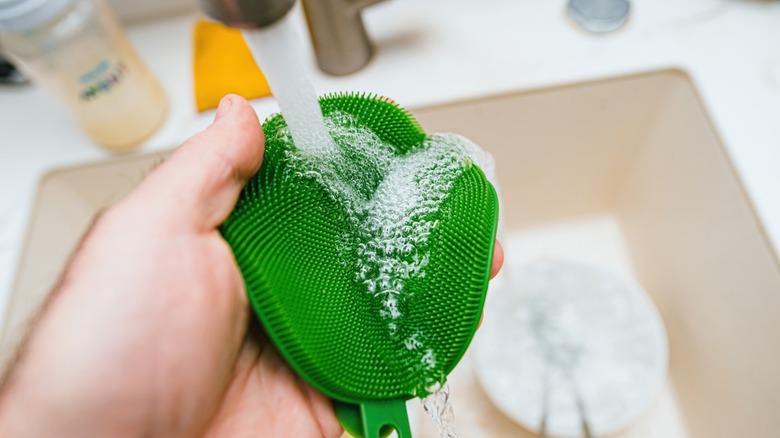 A green silicone sponge is washed under a faucet with soap over a sink