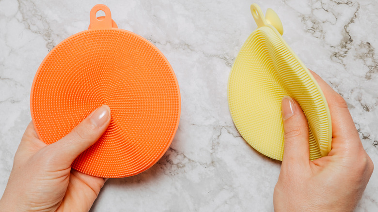 Two hands holding silicone sponges against a marble backdrop, one orange and one yellow
