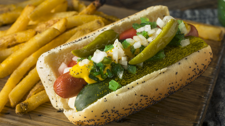 A Chicago-style hot dog with all the fixings on a wood board with a side of french fries