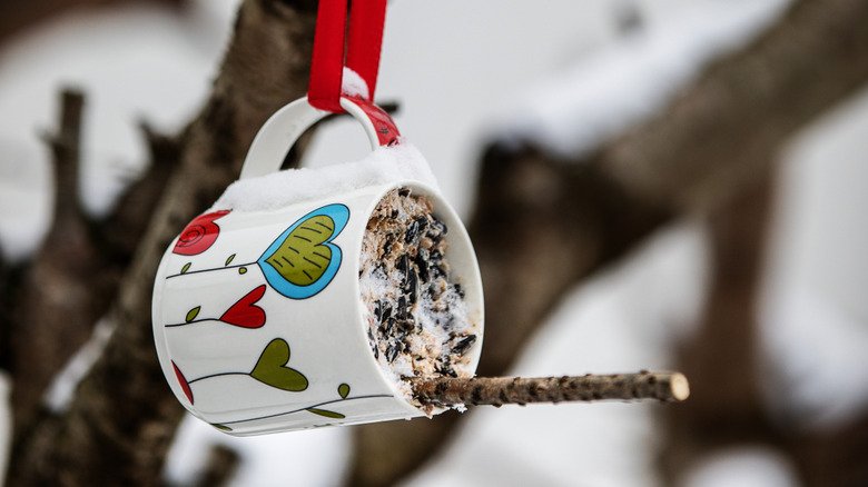 A coffee mug bird feeder hung by a ribbon with a stick perch