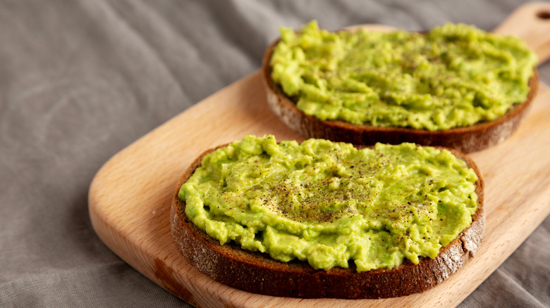 Two slices of bread topped with smashed avocado, displayed on a cutting board.