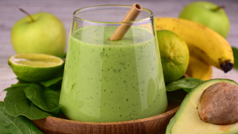 Green smoothie in short glass surrounded by spinach, green apples, avocados, and bananas
