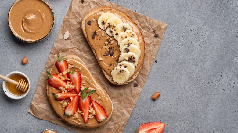 Slice of toast with peanut butter and banana next to slice of toast with peanut butter and strawberries next to bowls of honey and peanut butter