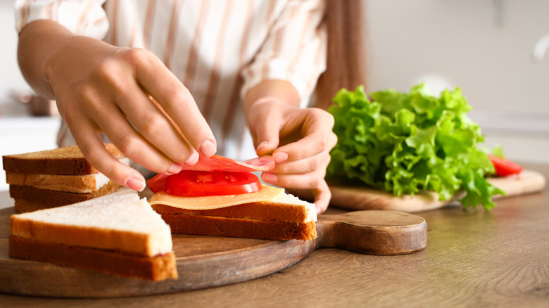 Someone making a sandwich, stacking meat and tomatoes on white bread, greens sitting on the counter beside it