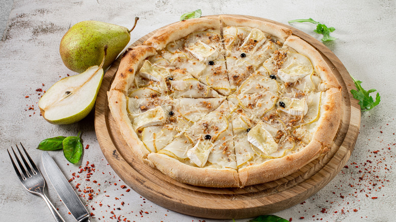 A brie and pear pizza sliced into triangles on a wooden pizza board with fresh pears, basil, arugula, and red pepper flakes sprinkled around the perimiter on the countertop