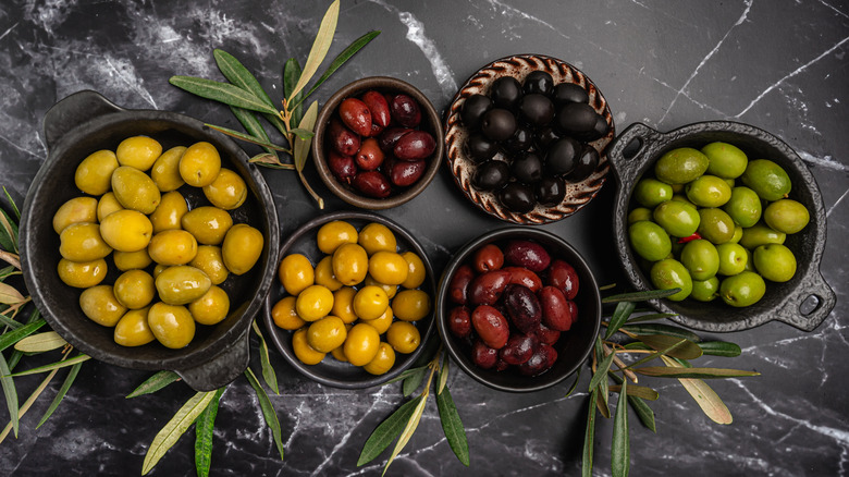 A black marble countertop with six small bowls with various types of olives