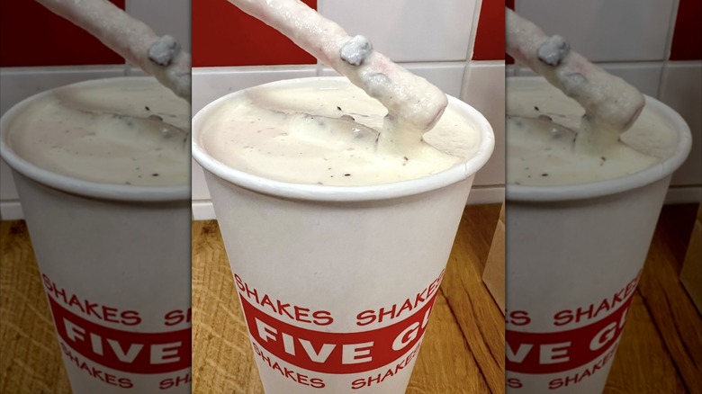 Five Guys milkshake in white paper cup with bacon bits on lifted straw, on wooden table facing tiled wall, with mirror borders