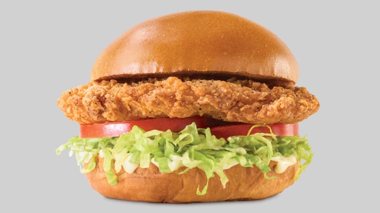 A crispy golden-fried chicken breast cutlet is served on a hamburger bun topped with a white-orange sauce, lettuce, and tomato