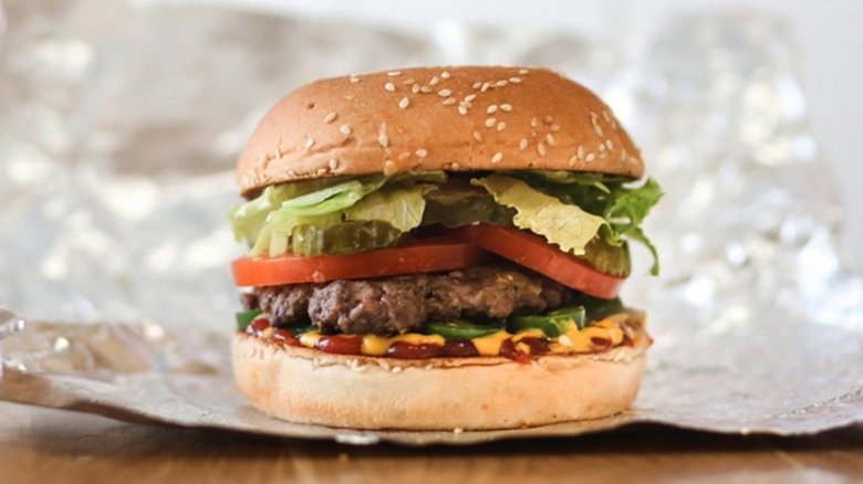 A single burger patty rests on a sesame seed hamburger bun topped with ketchup, mustard, jalapenos, pickles, and lettuce, on a single sheet of aluminum foil
