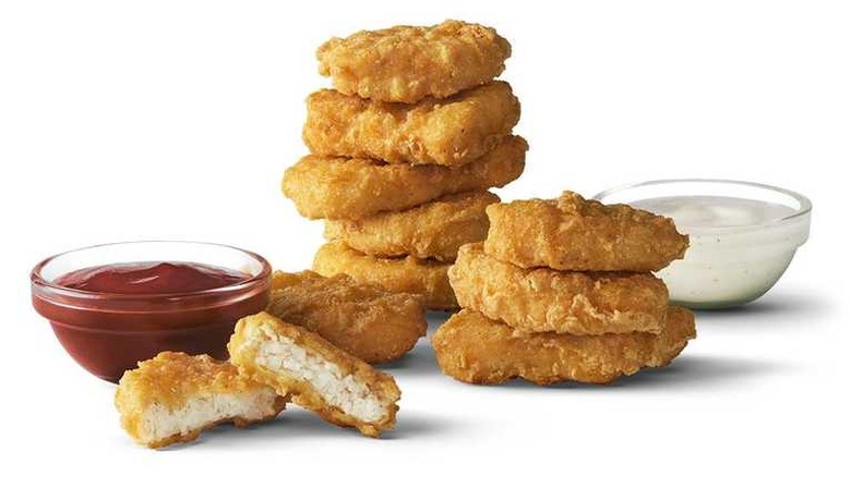 A small dipping cup filled with red ketchup sits next to piles of 10 stacked, golden fried chicken nuggets. Behind the nuggets is another dipping sauce cup filled with a white sauce.