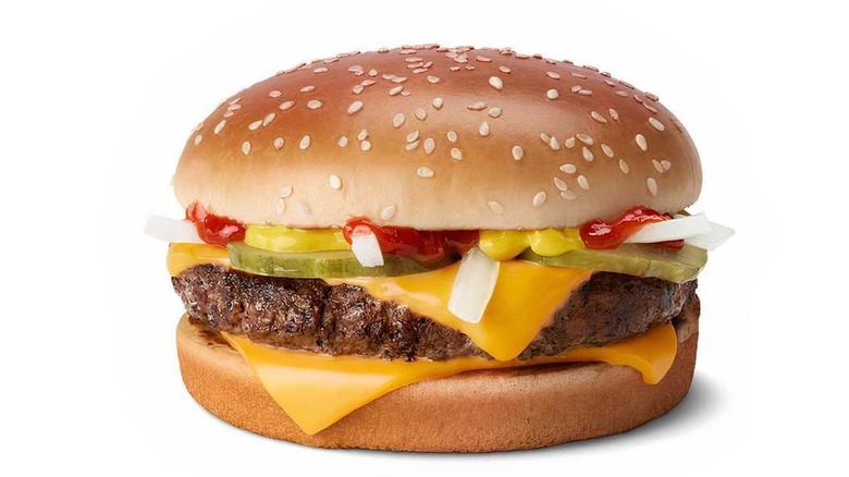 A closeup of a cheeseburger on a sesame seed bun. The yellow American cheese is melted and surrounds the single quarter-pound beef patty, which is topped with pickles, diced onions, ketchup, and mustard