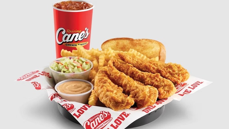 A Raising Cane's branded red-fountain drink cup is filled with a dark brown soda and ice to the brim. In front of the fountain drink is a basket lined with Raising Cane's branded logos, filled with a small serving of coleslaw, crinkle-cut french fries, Texas toast, four golden fried chicken fingers, and a cup of Raising Cane's sauce, which is orangey-red in appearance, on a white background.