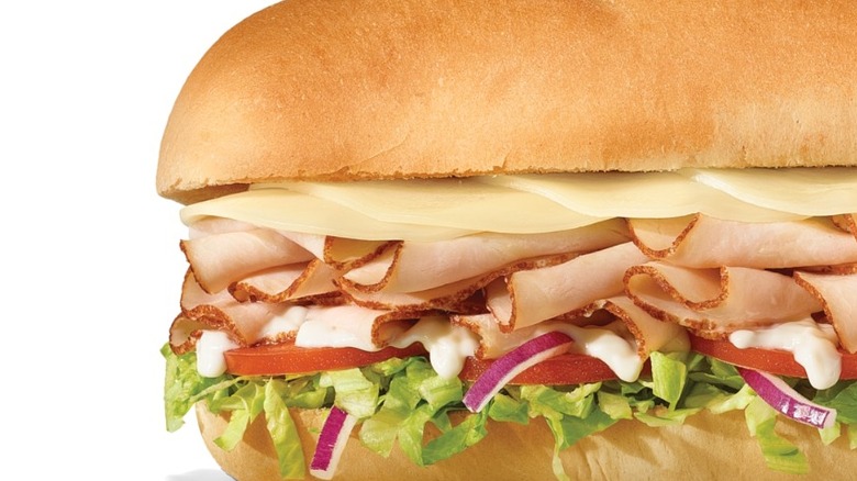 A baguette is sliced in half and topped with lettuce, red onion, tomato, mayo, deli-sliced turkey, and cheese