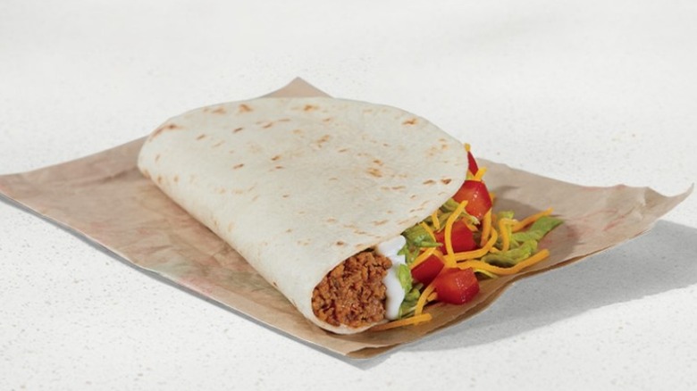 A single soft flour tortilla is filled with ground beef, sour cream, lettuce, tomato, and cheese, folded in half, and placed on its side on a countertop on top of a brown paper wrapper