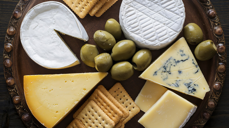 a variety of cheeses on a dark wood platter with olives and crackers