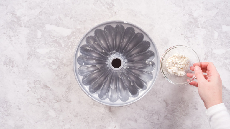 A greased bundt pan with a hand and a bowl of flour next to it