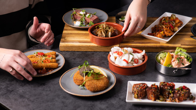 An array of small plates and bowls holding various tapas, a person's hand setting one down, and another pair of hands selecting a croquette of some sort