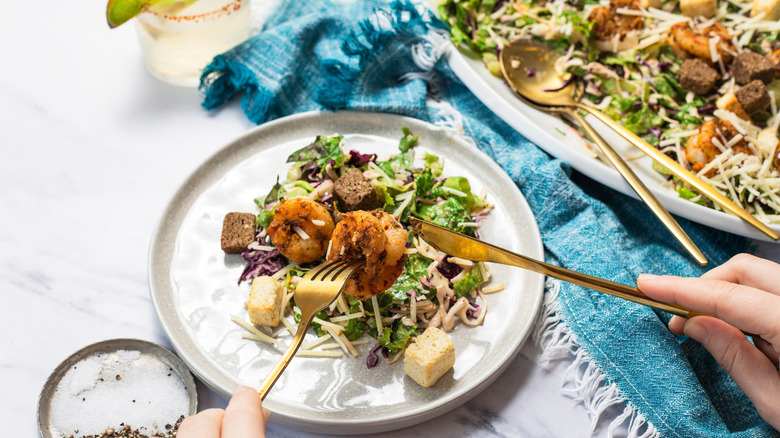 Lifting a shrimp off a plate with serving of Baja shrimp Caesar salad alongside margarita and platter.