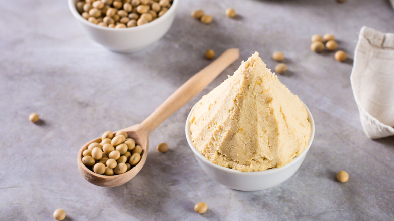 White miso in a bowl with a wooden spoon full of soybeans near it and another bowl of soybeans in the background.