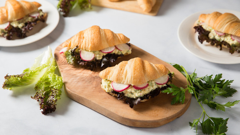 Two egg salad croissant sandwiches on a wooden board and others on plates