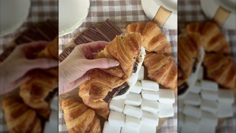 Hand holding a s'mores-stuffed croissant over a spread of croissants, marshmallows, and chocolate