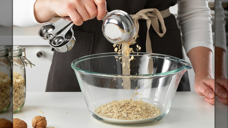 A baker measuring oats into a bowl with a metal measuring cup