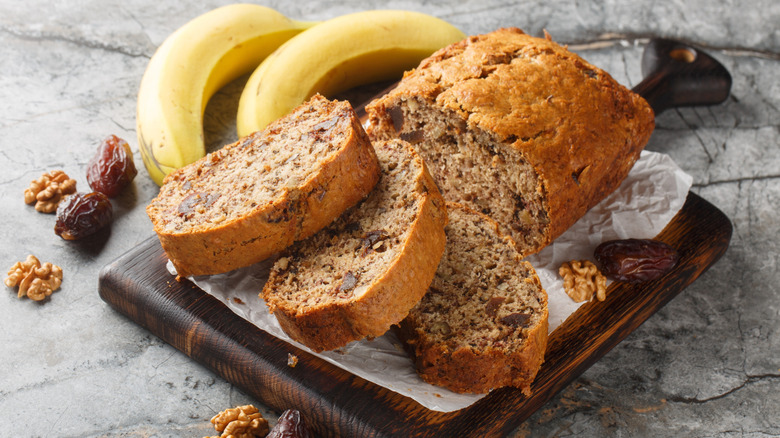 Sliced loaf of banana bread on wooden board next to whole bananas and pecans