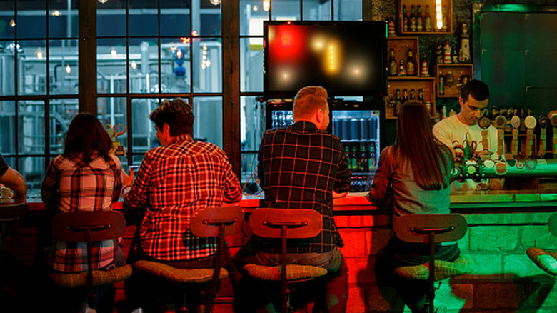 A number of bar patrons sitting at a bar viewed from behind, while a bartender stands behind the bar