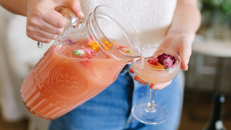 Person pouring pink cocktail topped with flowers from pitcher into coupe glass