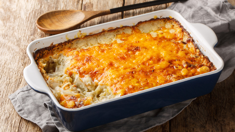 A cheesy, baked casserole in a dish sits on a napkin on a wooden table with a wooden spoon next to it