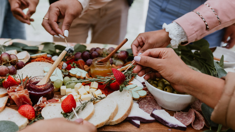 People's hands dice into a charcuterie board with cheese, bread, jam, honey, olives, grapes, strawberries, and cured meat