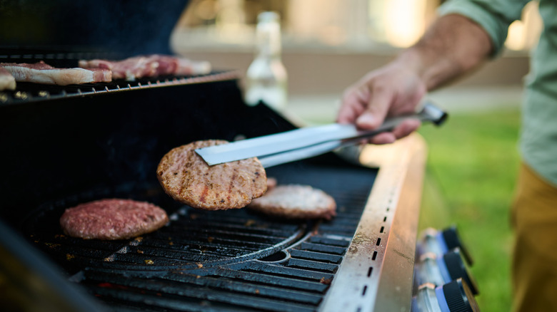 A person holds a burger patty over a grill with tongs