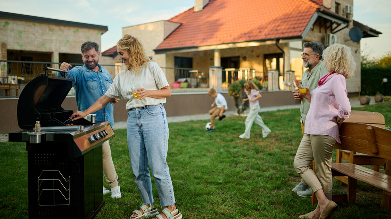 an affluent suburban back yard, with a younger Caucasian couple and a middle-aged Caucasian couple gathered around a gas grill, while children play soccer in the background