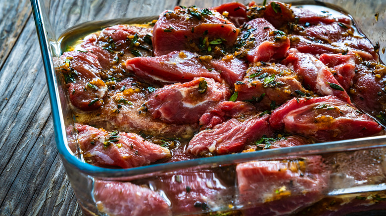 a glass baking dish, filled with marinating raw meat, sitting out on a rough, rustic wooden surface
