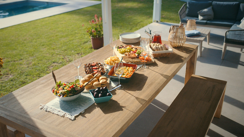 a tray of sliders and several colorful side dishes on a long wooden table with bench seating, on a concrete paio