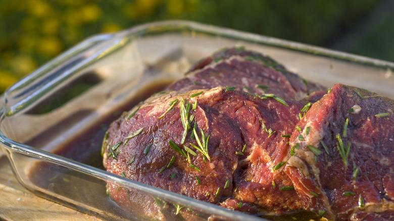ribeye steaks stacked in a glass baking dish, with a dark-colored marinade and coarsely chopped fresh rosemary