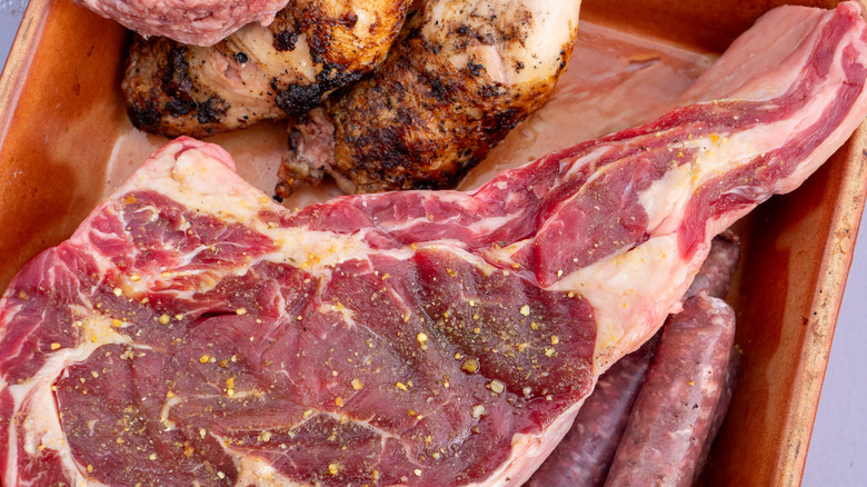 a raw, heavily marbled ribeye steak, a pair of uncooked sausages, and the edge of a raw hamburger all sharing a container with two pieces of already-cooked food