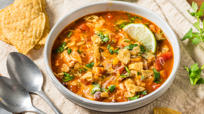 Bowl of chicken tortilla soup next to herbs, tortilla chips, and spoons