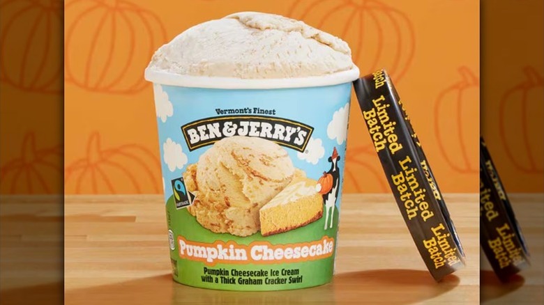 A beauty shot of a pint of Ben & Jerry's Pumpkin Cheesecake Ice Cream sitting on a wooden table with a pumpkin illustrated background