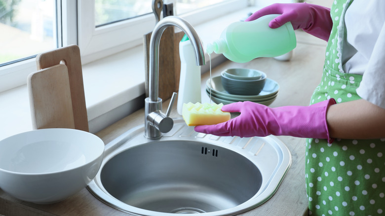 A woman wearing an apron and pink gloves squeezing dishwashing liquid onto a sponge at a kitchen sink