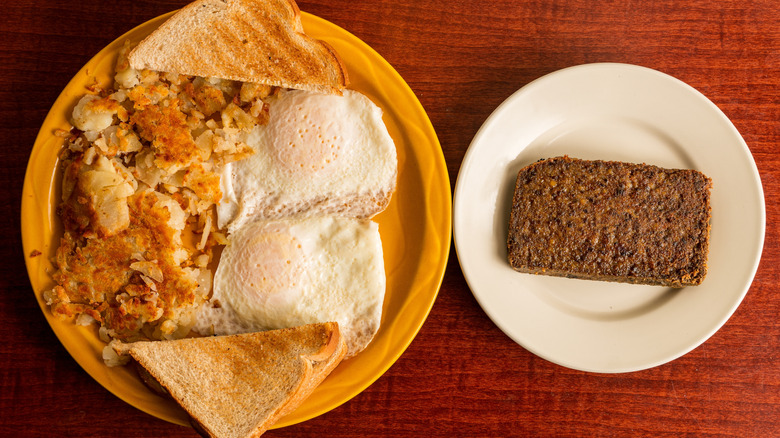 A plate with eggs, potatoes, and toast sits next to a plate with a piece of scrapple on it.