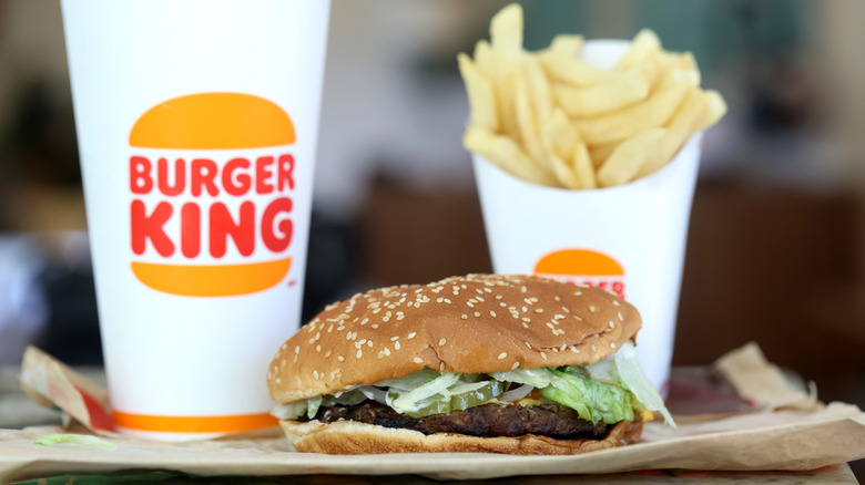 A Burger King Whopper on a paper bag with a soft drink and container of french fries behind it
