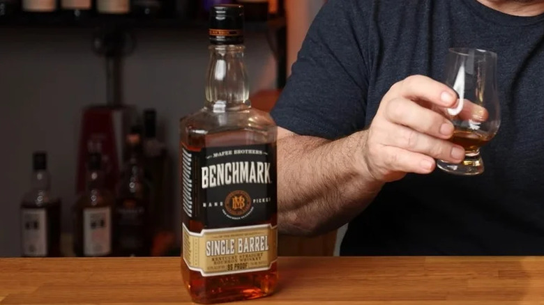 Someone holding a glass of Benchmark Single Barrel with the bottle on the bar top in front of them