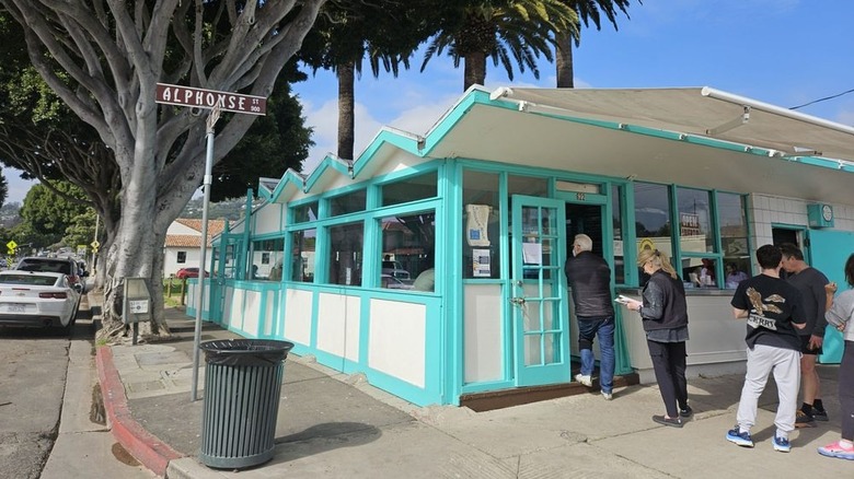 Customers standing in line at a one-story building on a tree-lined street