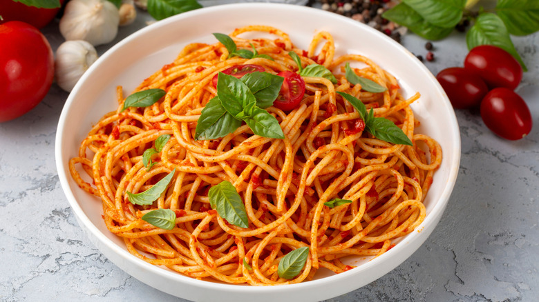 A white bowl of spaghetti in tomato sauce garnished with fresh basil leaves, surrounded by tomatoes, garlic bulbs, basil leaves, and peppercorns