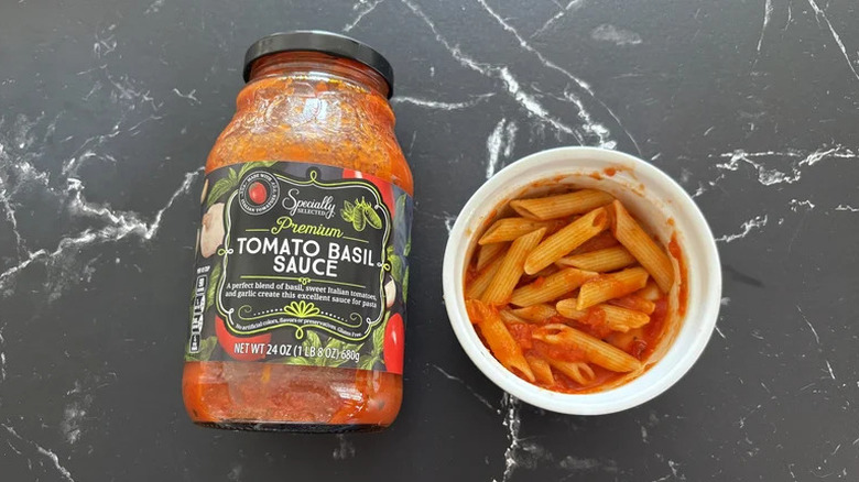 A jar of Specially Selected Premium Tomato Basil Sauce on a marble countertop, next to a bowl of cooked penne pasta dressed with the sauce