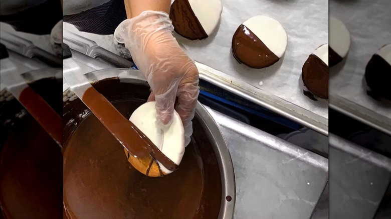 Hand holding a black and white cookie over a pot with a knife, applying the chocolate icing