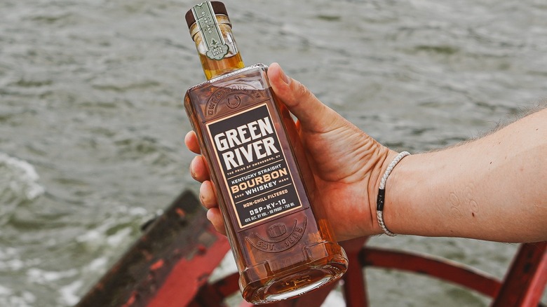 A person holding up a bottle of Green River Bourbon to camera with a background of open water