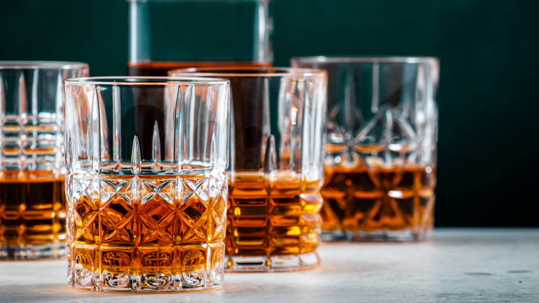 Four filled glasses of whiskey on a table, with a bottle shown in the blurred background